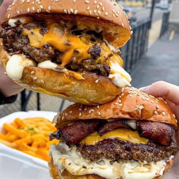 Two hands holding stacked Crazy Good Kitchen burgers with melted cheese, bacon, and fries in the background.