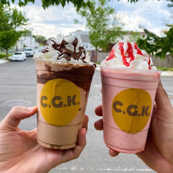 Two hands holding Crazy Good Kitchen milkshakes, one chocolate and one strawberry, topped with whipped cream.