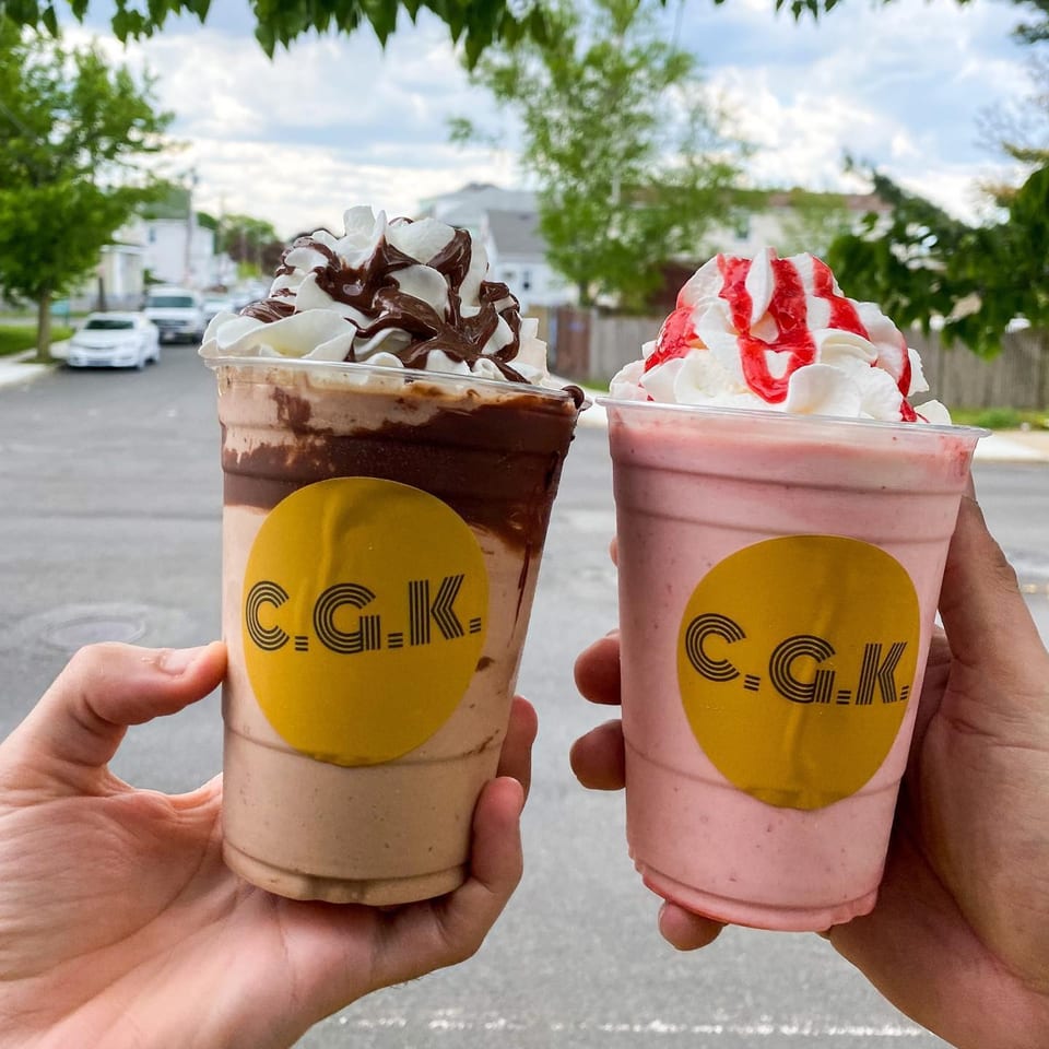 Two hands holding Crazy Good Kitchen milkshakes, one chocolate and one strawberry, topped with whipped cream.