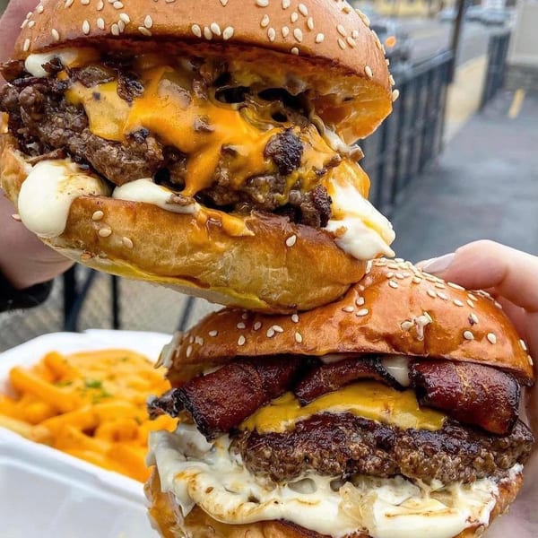 Two hands hold up two Crazy Good Kitchen burgers, one with bacon, over a container of fries.
